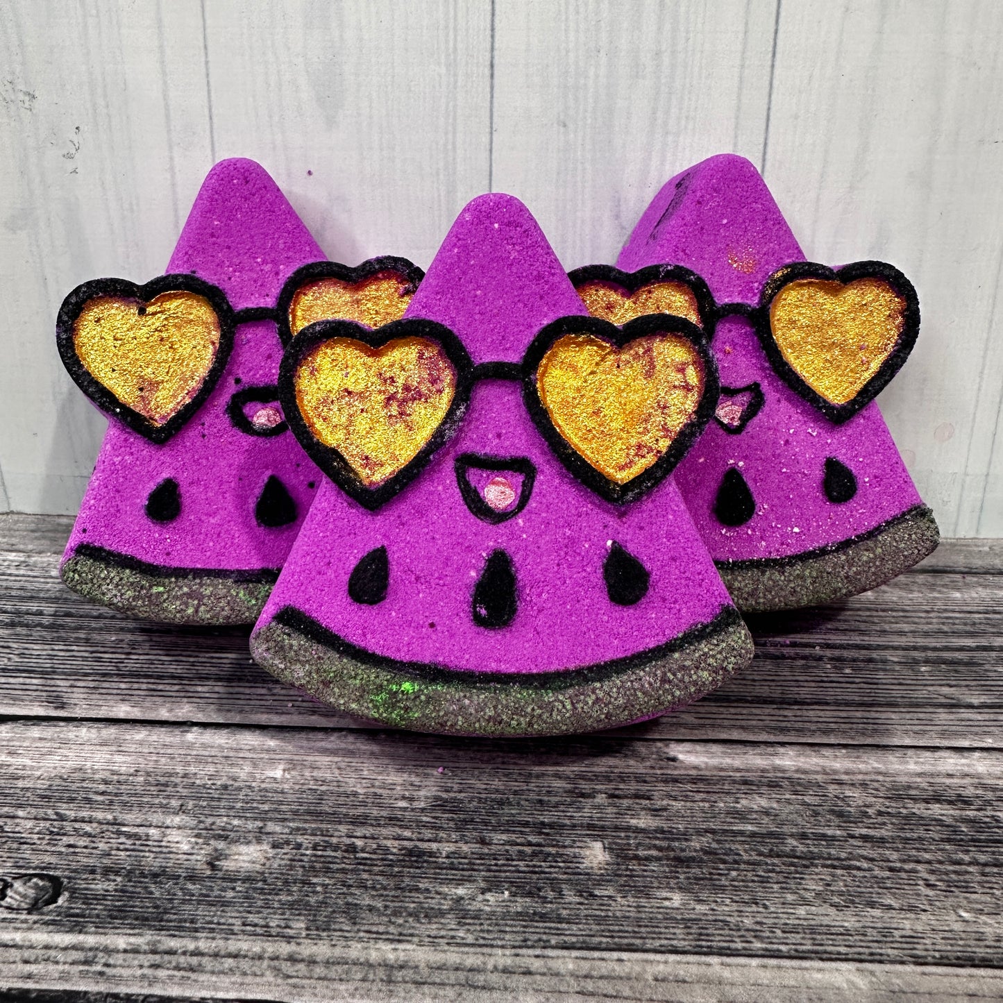 A bright pink triangle shaped bath bomb made to look like a watermelon slice, with gold painted love heart sunglasses